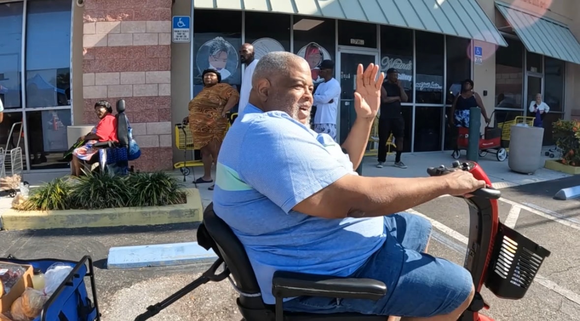 Community neighbor waving from a mobility scooter at the Saturday food distribution at Tangerine Plaza